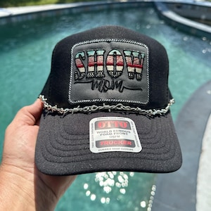 May include: Black trucker hat with a barbed wire brim. The hat features a patch with the words "SHOW MOM" in a patriotic red, white, and blue design. The hat has a snap closure and a foam front.
