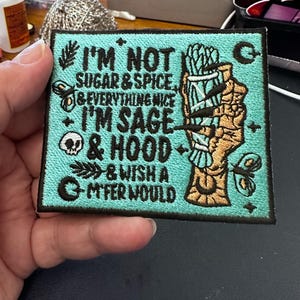 May include: A turquoise blue embroidered patch with black text that reads "I'm not sugar & spice & everything nice I'm sage & hood & wish a m'fer would". The patch features a hand holding a bundle of sage and a crescent moon.