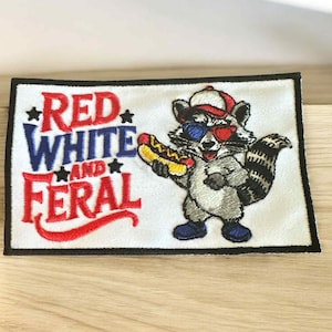 May include: Embroidered patch with the text "RED WHITE AND FERAL". The patch features a raccoon wearing a hat and 3D glasses, holding a hot dog. The patch has a black border and is on a white background.