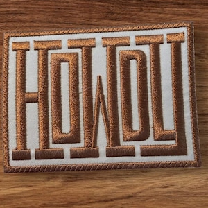 Pode incluir: Patch bordado com a palavra "HOWL" em letras grandes e em bloco. O texto é de cor castanho metálico, costurado sobre um fundo branco. O patch tem uma borda castanha e é de forma retangular.