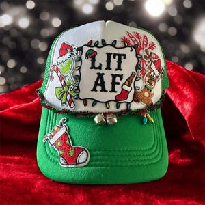 Puede incluir: Gorra de camionero navideña verde y blanca con el texto "LIT AF" en un parche blanco. El sombrero presenta adornos festivos como un reno, un calcetín y un bastón de caramelo. También se adjuntan pequeñas campanas y luces decorativas.