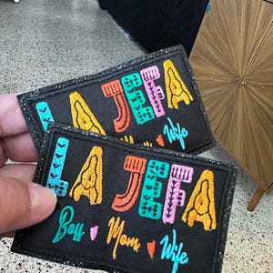 May include: Two black fabric patches with colorful embroidered text. The top patch reads "LA JEFA Wife". The bottom patch reads "LA JEFA Boss Mom Wife".