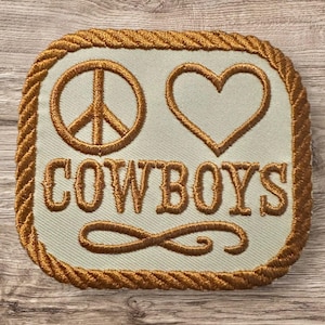 May include: Embroidered patch with a light green background, featuring a peace sign, a heart, and the word "COWBOYS" in a serif font. The patch is bordered with a gold rope-like trim and a decorative flourish.