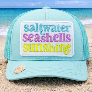 May include: A light blue trucker hat with a mesh back and a curved brim. The hat features a white patch with the words "saltwater seashells sunshine" in turquoise, purple, and yellow. A small seashell is attached to the brim.