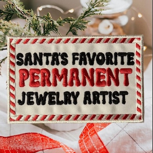 May include: Embroidered patch with the text "SANTA'S FAVORITE PERMANENT JEWELRY ARTIST" in black and red lettering. The patch has a cream-colored background with a red and white striped border. Festive holiday theme.