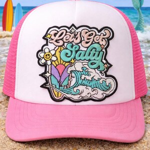 May include: Pink and white trucker hat with a patch that says "Let's Get Salty." The patch features a flower, a wave, and a person surfing. The hat has a pink brim and mesh back. Surfboards and a mermaid tail are in the background.