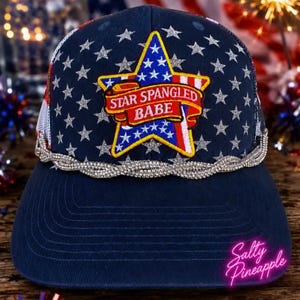 May include: A navy blue trucker hat featuring a silver glitter star pattern and a central patch shaped like a star with a red banner reading STAR SPANGLED BABE. The hat is accented with a braided rhinestone trim along the base of the front panel.