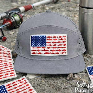 Può includere: Un cappellino grigio a tesa piatta con una toppa raffigurante una bandiera americana, dove le strisce sono sostituite da sagome di pesci rossi. Nell'immagine sono visibili anche attrezzatura da pesca e altre toppe con bandiere.