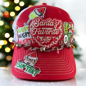 Peut inclure: Casquette de baseball rouge avec un dos en filet blanc, ornée d'écussons et de texte sur le thème de Noël. Les écussons incluent le Père Noël, les mots "Santa's Favorite Ho" et "I SAW THAT". La casquette est ornée d'une guirlande de petites lumières et de cloches.