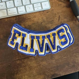 May include: A blue, white, and gold embroidered patch with the word "FLIVVS" in bold letters. The patch has a curved shape and is likely designed to be sewn onto clothing or accessories. The patch is on a wooden surface.