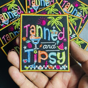 Puede incluir: Parche bordado con las palabras "Tanned and Tipsy" en letras coloridas. El diseño incluye un cóctel, palmeras, un sol y una rodaja de sandía. El parche tiene un fondo negro y un borde amarillo.