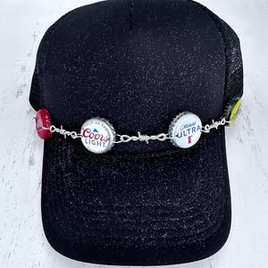 May include: Black trucker hat with a mesh back, decorated with bottle caps from Coors Light, Michelob Ultra, and another beer brand. The bottle caps are attached with a silver chain and barbed wire.
