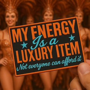 Puede incluir: Parche rectangular negro con texto naranja y turquesa que dice: "MY ENERGY Is a LUXURY ITEM. Not everyone can afford it." El parche se sostiene frente a un fondo borroso de personas con trajes de lentejuelas naranjas.