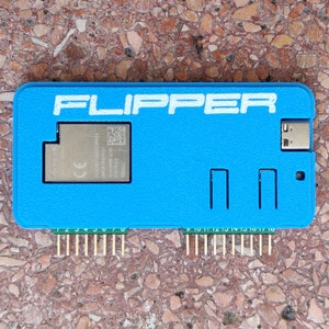 以下が含まれることがあります： 白い文字で「FLIPPER」と印刷された青いプラスチックケース。ケースには、CEマークとQRコードがついた小さな銀色のチップが入っています。ケースの下部には、金色のピンが18本あります。