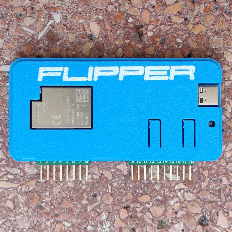 以下が含まれることがあります： 白い文字で「FLIPPER」と印刷された青いプラスチックケース。ケースには、CEマークとQRコードがついた小さな銀色のチップが入っています。ケースの下部には、金色のピンが18本あります。