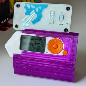 May include: A purple handheld electronic device with a white screen displaying a game. The device has a large orange button with directional arrows and the text "RESET" and "BOOT" on the top.