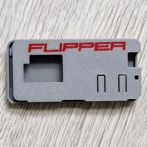 以下が含まれることがあります： 赤い文字で「FLIPPER」と書かれた灰色のプラスチックケース。ケースには長方形の切り抜きが3つあります。