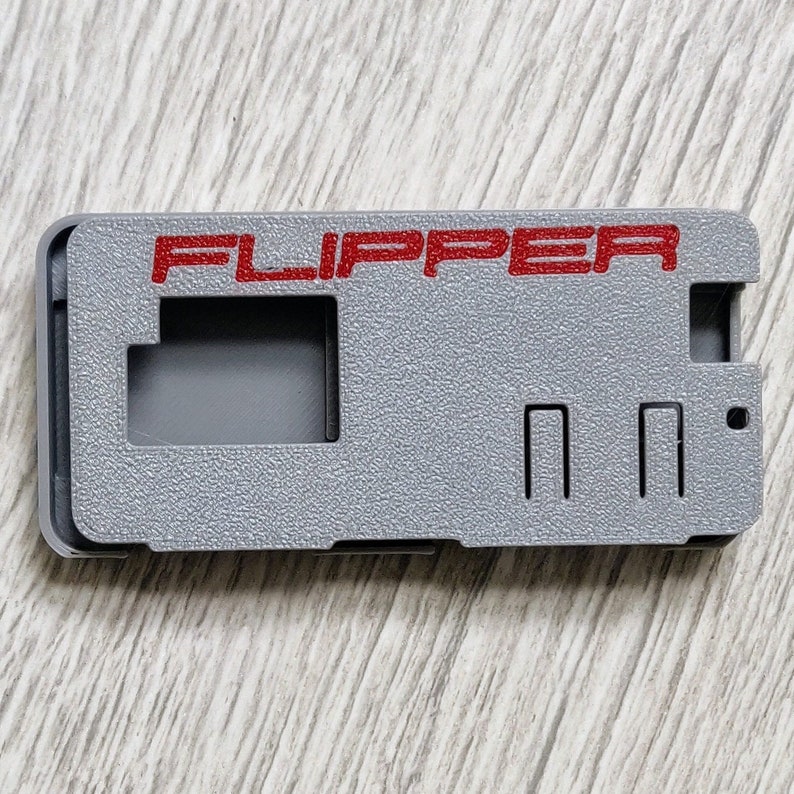 以下が含まれることがあります： 赤い文字で「FLIPPER」と書かれた灰色のプラスチックケース。ケースには長方形の切り抜きが3つあります。