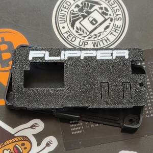 以下が含まれることがあります： 白い文字で「FLIPPER」と印刷された黒いプラスチック製のケース。ケースには長方形の開口部と右側に小さな長方形のスロットがあります。