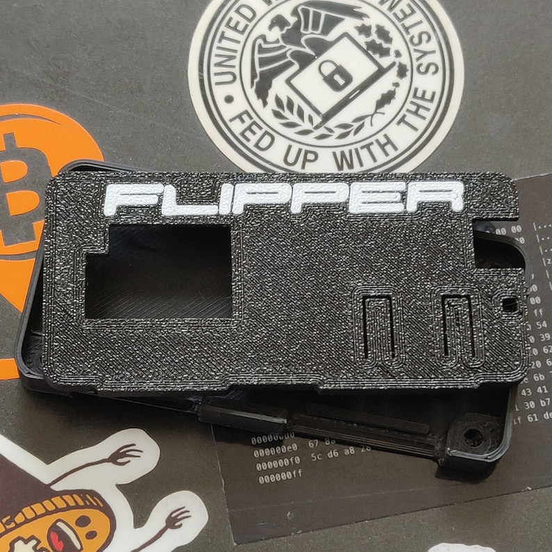 以下が含まれることがあります： 白い文字で「FLIPPER」と印刷された黒いプラスチック製のケース。ケースには長方形の開口部と右側に小さな長方形のスロットがあります。