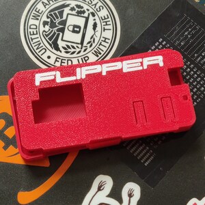 以下が含まれることがあります： 白い文字で「FLIPPER」と印刷された赤いプラスチック製のケース。ケースには長方形の切り抜きが2つあり、表面はテクスチャ加工されています。