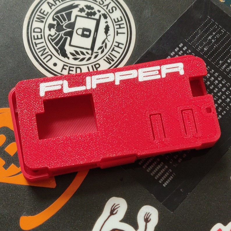 以下が含まれることがあります： 白い文字で「FLIPPER」と印刷された赤いプラスチック製のケース。ケースには長方形の切り抜きが2つあり、表面はテクスチャ加工されています。
