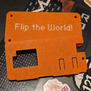 May include: Orange plastic case with the text "Flip the World!" printed in white. The case has a textured surface and several rectangular cutouts.