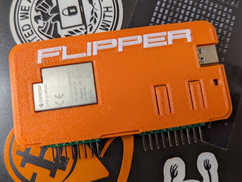 以下が含まれることがあります： 前面に白い文字で「FLIPPER」と印刷されたオレンジ色のプラスチック製のデバイス。デバイスには、前面に小さな金属チップと、底面にピンが並んでいます。