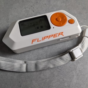 Pode incluir: Um dispositivo de mão branco e laranja com uma tela preta que exibe o texto "Flipper" e uma alça. O dispositivo possui um botão redondo laranja com um pequeno botão preto ao lado.