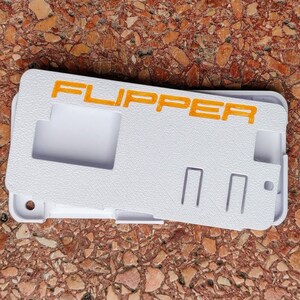以下が含まれることがあります： オレンジ色の文字で「FLIPPER」と印刷された白いプラスチックケース。ケースには長方形の切り抜きと2つのスロットがあります。