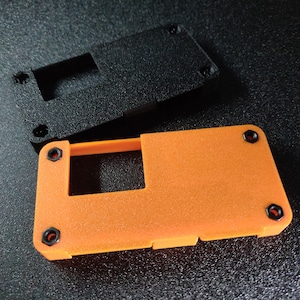 Op de afbeelding: Een oranje plastic behuizing met een zwart plastic deksel. De behuizing heeft een rechthoekige opening en verschillende kleine zwarte schroeven.