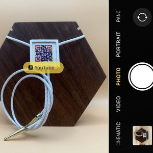 May include: White bolo tie with a gold-colored metal tip and a white square pendant with a black and white QR code and the text "YouTube". The bolo tie is displayed on a hexagonal wooden stand.