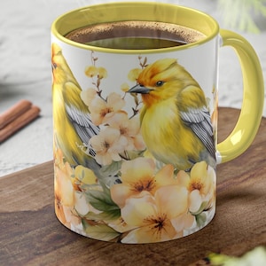May include: A yellow coffee mug with a yellow rim and handle features a watercolor illustration of two yellow birds perched on a branch with white and yellow flowers.