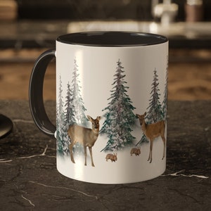 May include: A white ceramic mug with a black rim and handle. The mug features a wintery scene with two deer and three rabbits in a snowy forest.