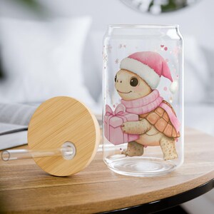 May include: A clear glass tumbler with a bamboo lid and straw. The tumbler features a cartoon turtle wearing a pink Santa hat and scarf, holding a pink gift. The design includes snowflakes and a white pom-pom.