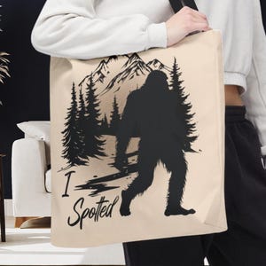 May include: Beige tote bag featuring a black silhouette of Bigfoot walking in a forest scene with mountains in the background. The text "I Spotted" is printed below the image. The bag has a black shoulder strap.