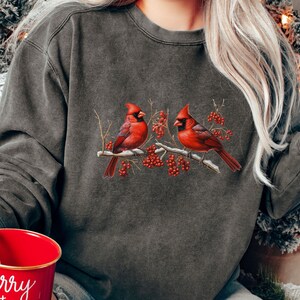 May include: A gray sweatshirt with a vintage look featuring a design of three red cardinals perched on a branch with red berries. The sweatshirt is worn by a person holding a red mug with the text "merry + bright" in white.
