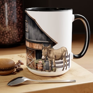 May include: White ceramic mug with a black rim and handle. The mug features a watercolor illustration of a donkey standing in front of a barn with a wooden door and a window. The donkey is brown and white and has a pink flower in its mouth.