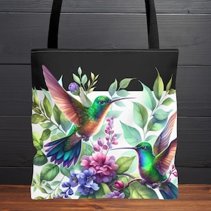 May include: Black tote bag featuring a vibrant watercolor design of two hummingbirds with green, blue, and orange plumage. The birds are surrounded by lush green foliage and colorful flowers, including pink and purple blooms. The bag has black handles.