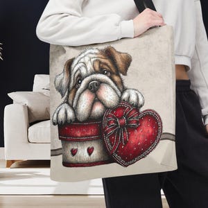 May include: Tote bag featuring a detailed illustration of a bulldog puppy peeking out of a gift box. The box is red with white accents and heart details, and the puppy is holding a red heart-shaped gift with a bow. The bag has a black strap.