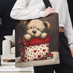 May include: A brown tote bag featuring a cute puppy peeking out of a red-trimmed gift box decorated with red hearts. A large red bow with white polka dots adorns the box. The bag has a black shoulder strap.