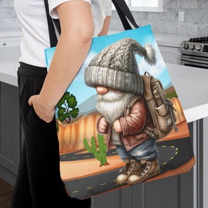 Op de afbeelding: Een blauwe tote bag met een cartoonillustratie van een kabouter die een grijze gebreide muts en een bruin jack draagt. De kabouter draagt een bruine rugzak en loopt op een zandweg met een cactus en een groene struik op de achtergrond.