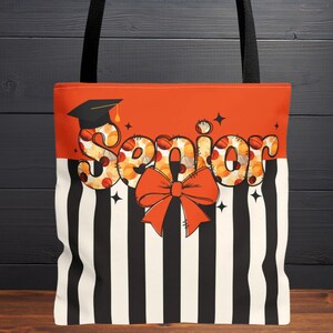 Pode incluir: Uma sacola com alça preta. A seção superior é laranja e apresenta a palavra "Senior" em uma fonte decorativa. A sacola tem um grande laço laranja e uma seção inferior listrada em preto e branco. Um chapéu de formatura está à esquerda.