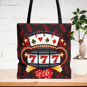 May include: Black canvas tote bag with a casino themed design. The design features a slot machine with the numbers 777, playing cards, and dice. The background is a black and white pattern of dollar signs.