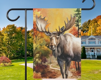 May include: A garden flag featuring a watercolour illustration of a brown moose standing in a woodland setting. The background depicts autumnal foliage in shades of orange, yellow, and red.