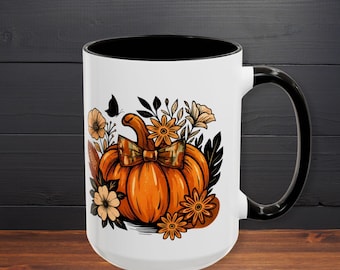May include: White ceramic mug with a black interior and handle. The mug features an orange pumpkin design with a plaid bow, surrounded by flowers and a butterfly. The mug is on a wooden surface.