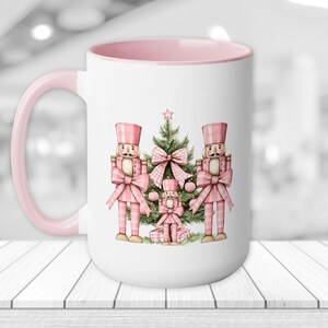 May include: White ceramic mug with a pink rim and handle. The mug features a watercolor illustration of three pink and white nutcracker figures standing in front of a green Christmas tree with pink and white bows.