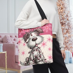 May include: A pink and white tote bag featuring a cartoon dog wrapped in Christmas lights. The top section is pink with snowflake patterns, and the bottom section is off-white with pink snowflakes. The bag has a black strap.