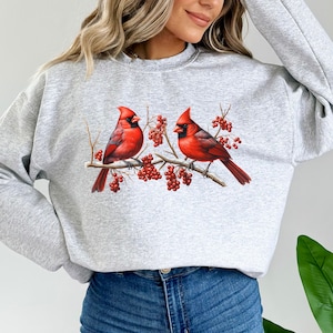 May include: A gray sweatshirt with a graphic of two red cardinals perched on a branch with red berries. The cardinals are facing each other and have black eyes and beaks.