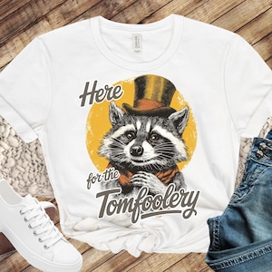 May include: White t-shirt featuring a raccoon wearing a top hat and scarf, set against a yellow circle. The text "Here for the Tomfoolery" is printed below the raccoon illustration. The shirt is paired with white sneakers and blue jeans.